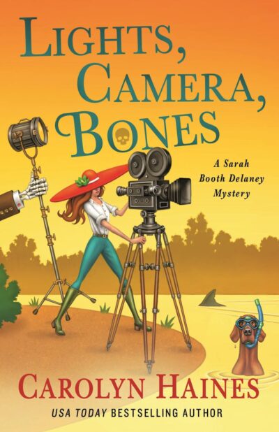 The cover of Lights, Camera, Bones by Carolyn Haines is an illustration of a woman in a big red hat operating a reel-to-reel camera, a skeleton hand holding a stage light, and a dog wearing a snorkel mask